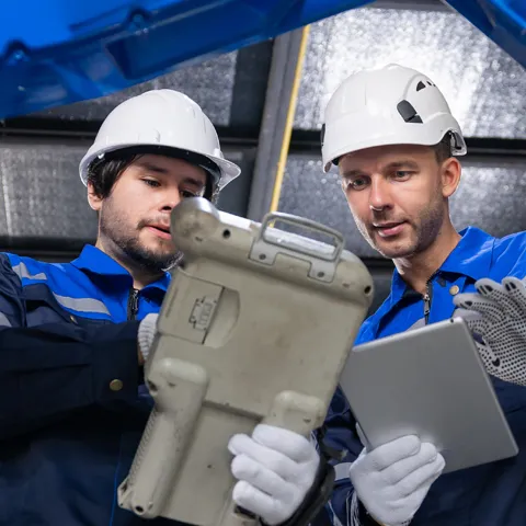 Engineers wearing hardhats using digital control pendant to inspect smart robotic arm system in industrial factory.